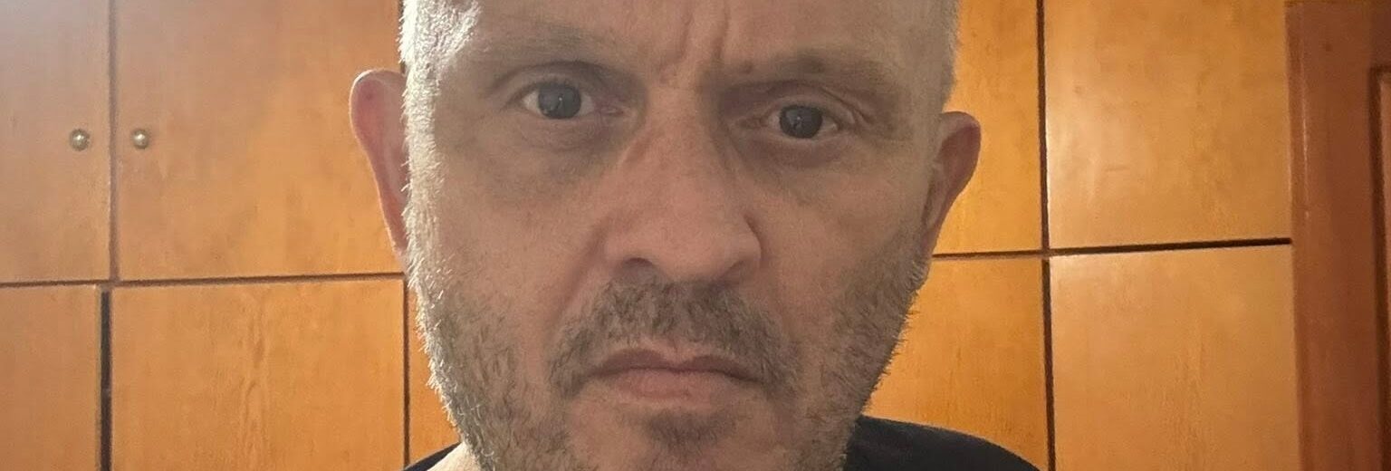 Άνδρας με γκρίζα μαλλιά και γένια, σοβαρό ύφος, φοράει μαύρο μπλουζ…