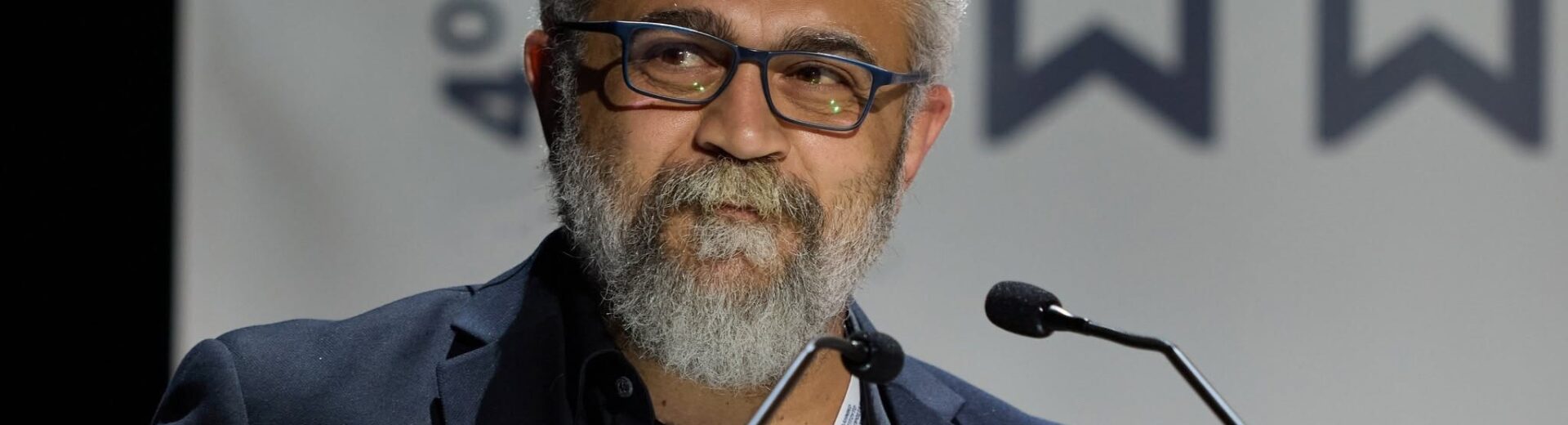 Άνδρας με γκρίζα γενειάδα και γυαλιά μιλάει σε μικρόφωνο σε εκδήλω…