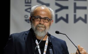 Άνδρας με γκρίζα γενειάδα και γυαλιά μιλάει σε μικρόφωνο σε εκδήλω…
