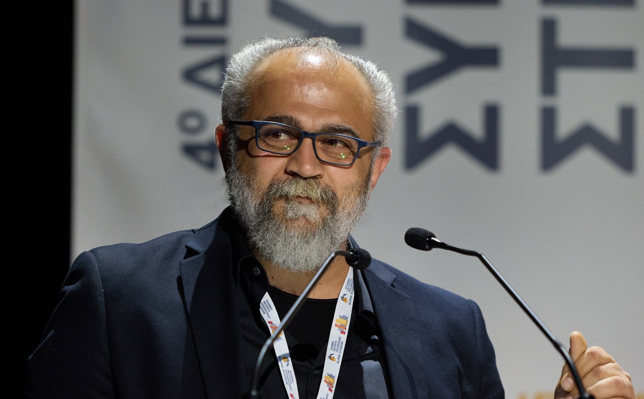 Άνδρας με γκρίζα γενειάδα και γυαλιά μιλάει σε μικρόφωνο σε εκδήλω…