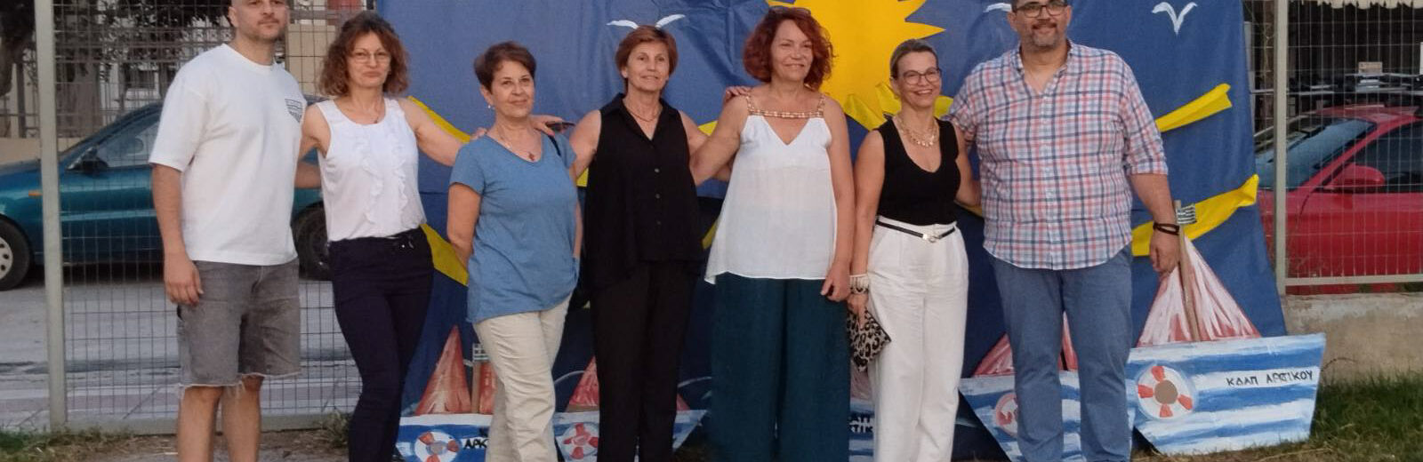 Επτά ενήλικες ποζάρουν μπροστά σε ένα μπλε καλοκαιρινό πανό με ήλι…