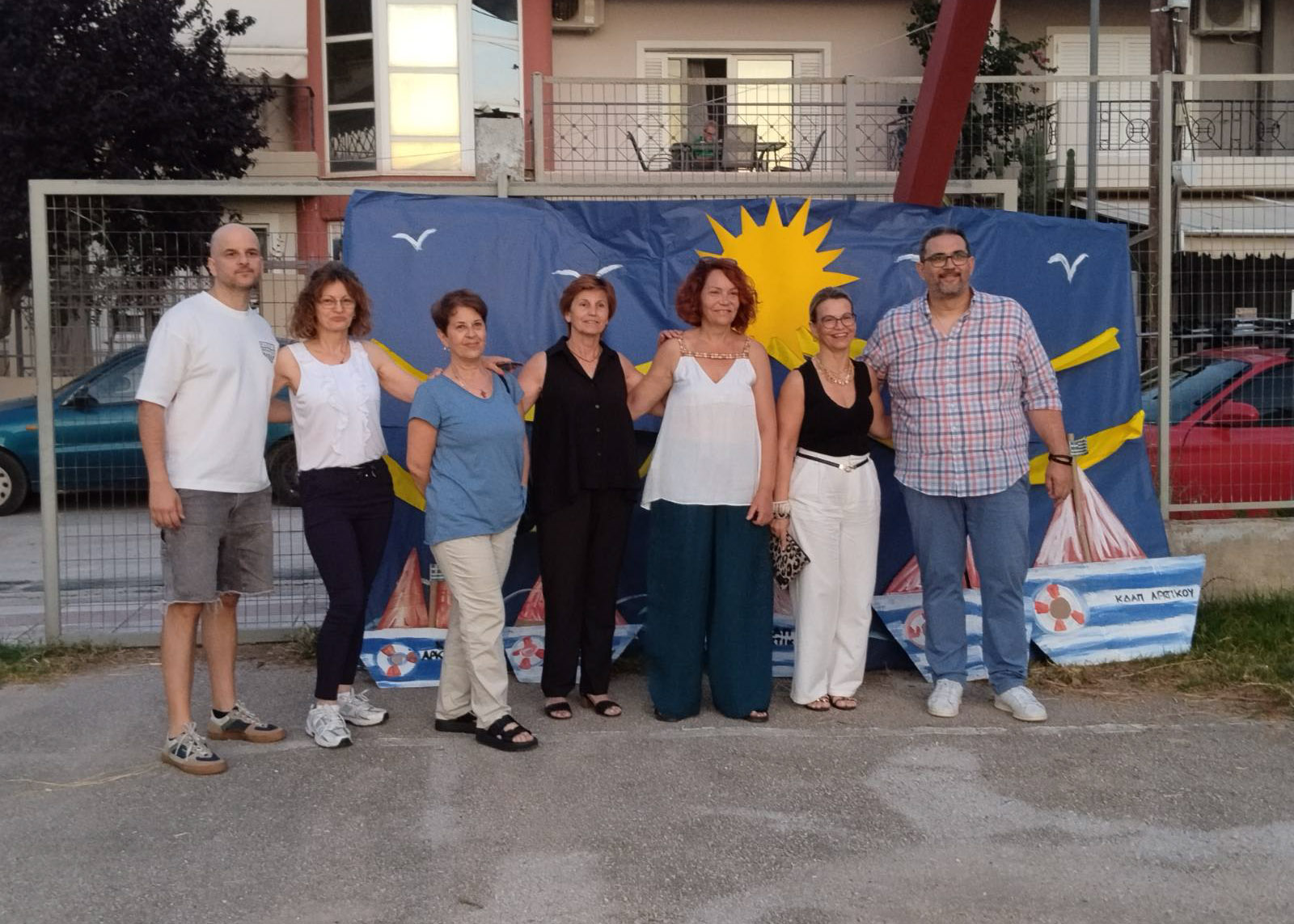 Επτά ενήλικες ποζάρουν μπροστά σε ένα μπλε καλοκαιρινό πανό με ήλι…