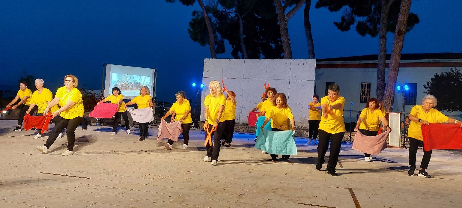 Τα ΚΑΠΗ Δήμου Αιγιαλείας στο Υπαίθριο Θέατρο ΦΩΤΟ 4 Γυναίκες από τα ΚΑΠΗ Αιγιαλείας με κίτρινες μπλούζες χορεύουν με π…