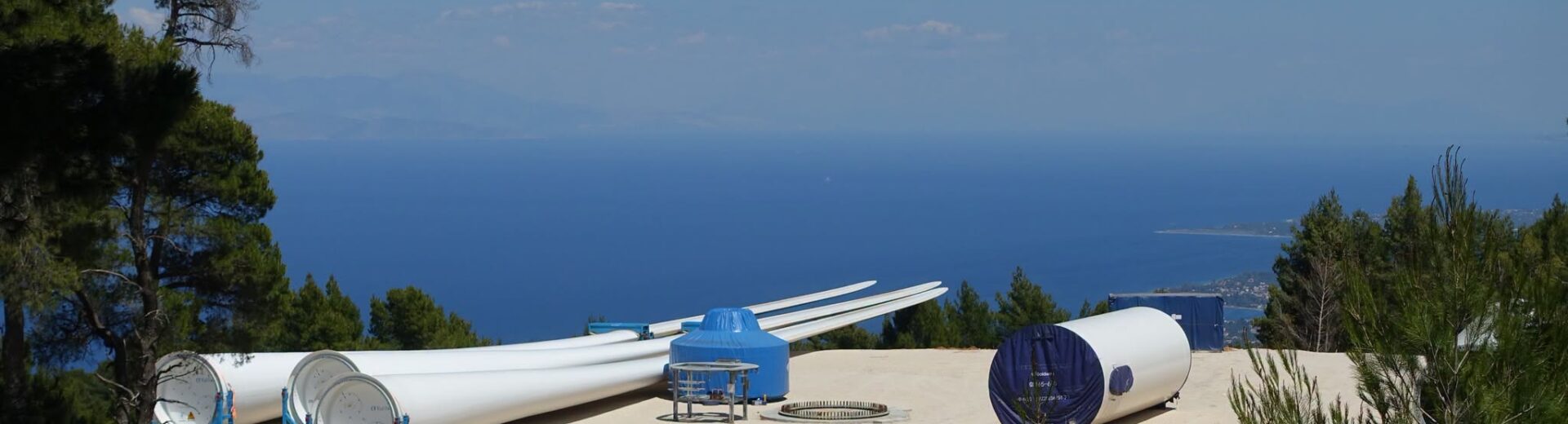 Τμήματα ανεμογεννητριών σε εργοτάξιο βουνού με θέα τη θάλασσα.