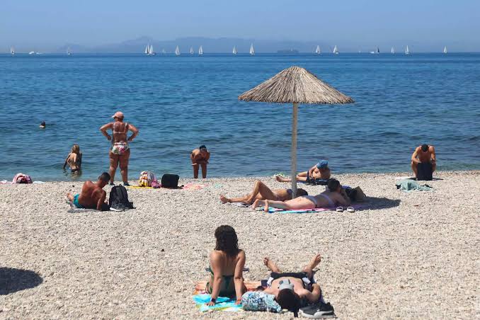 Άνθρωποι κάνουν ηλιοθεραπεία και κολυμπούν σε παραλία με βότσαλα, …