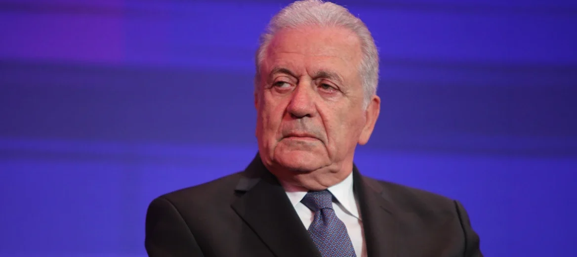 Ο Δημήτρης Αβραμόπουλος με κοστούμι και γραβάτα, κοιτάζει με σοβαρ…
