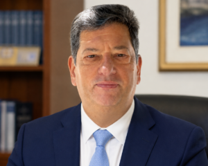Άνδρας με μπλε κοστούμι και γραβάτα ποζάρει σε γραφείο.