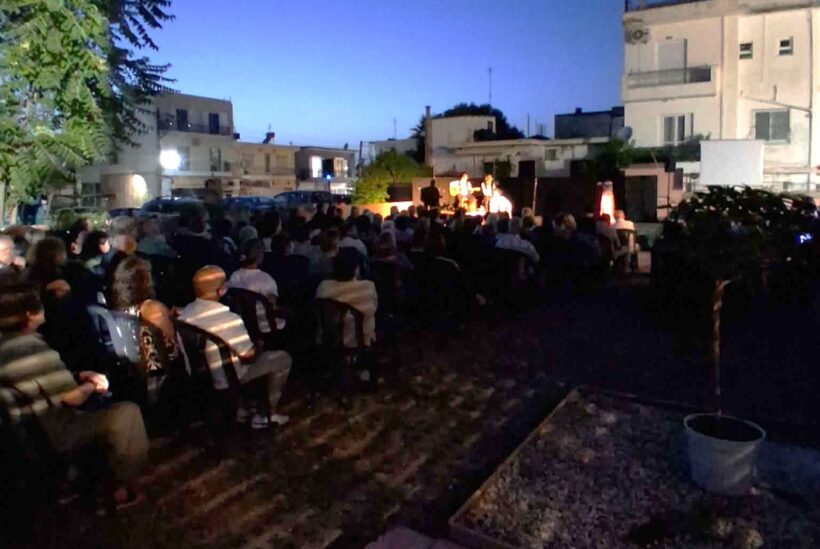 Κοινό παρακολουθεί μουσική εκδήλωση σε εξωτερικό χώρο το βράδυ, με…