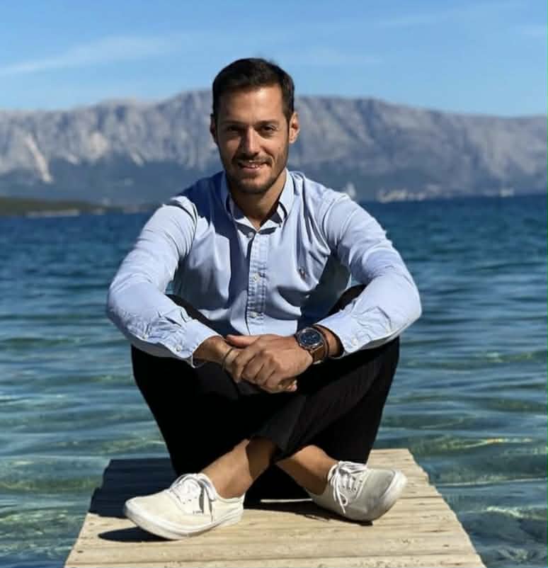 Νέος άνδρας χαμογελά καθισμένος σε ξύλινη προβλήτα με θάλασσα και …
