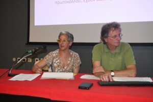 Μια γυναίκα μιλάει σε μικρόφωνο δίπλα σε έναν άνδρα, σε εκδήλωση πα…