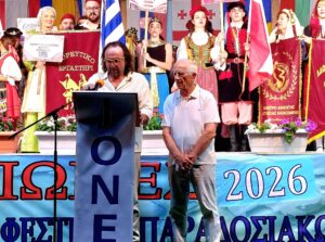 Δύο άνδρες σε βήμα, με άτομα με παραδοσιακές ενδυμασίες και σημαίες πίσω τους στο 20ο Παγκόσμιο Φεστιβάλ Παραδοσιακών Χορώ…