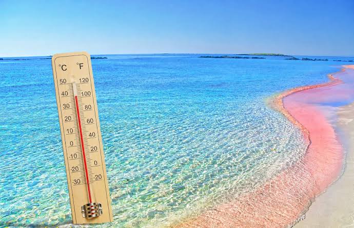 Θερμόμετρο σε παραλία με ροζ άμμο και γαλάζια νερά, δείχνει υψηλή θ…