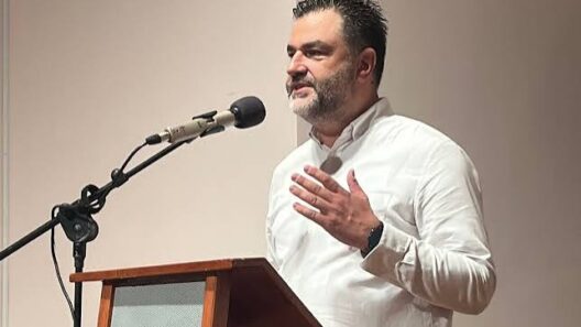 Άνδρας με γενειάδα και λευκό πουκάμισο μιλάει σε μικρόφωνο από βήμ…