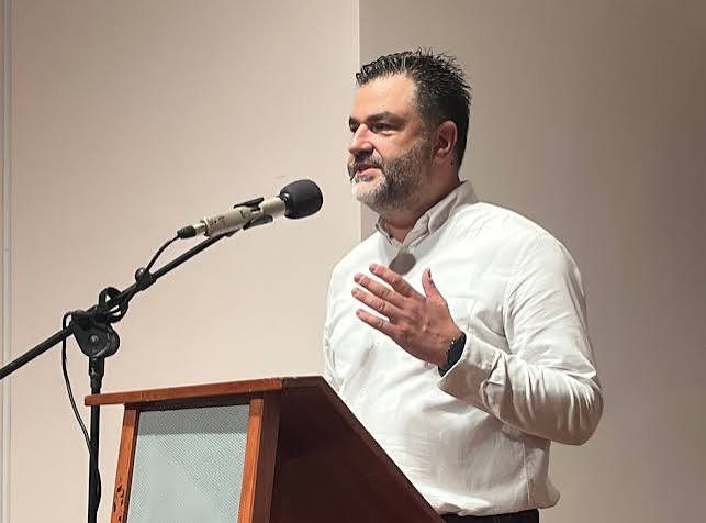 Άνδρας με γενειάδα και λευκό πουκάμισο μιλάει σε μικρόφωνο από βήμ…
