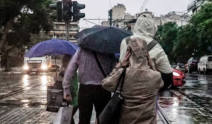 Άνθρωποι με ομπρέλες περπατούν σε βροχερό δρόμο πόλης.