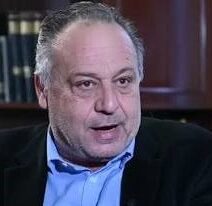 Άνδρας με γκρίζα μαλλιά και μπλε πουκάμισο μιλάει, με βιβλία στο φό…