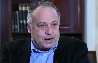 Άνδρας με γκρίζα μαλλιά και μπλε πουκάμισο μιλάει, με βιβλία στο φό…