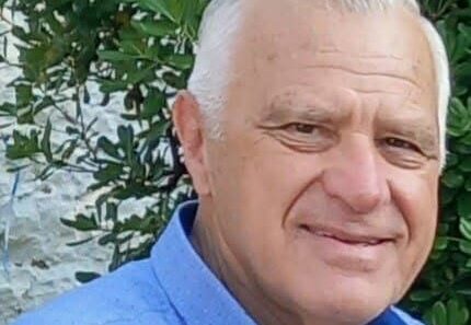 Ηλικιωμένος άνδρας με γκρίζα μαλλιά και μπλε πουκάμισο χαμογελά.