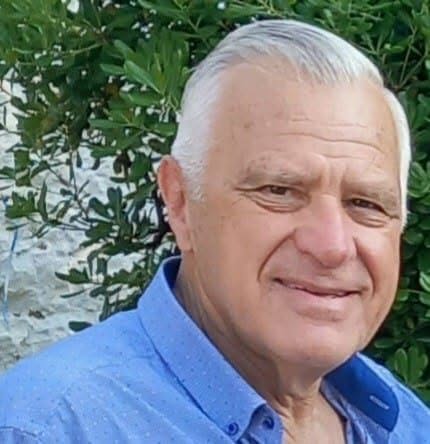Ηλικιωμένος άνδρας με γκρίζα μαλλιά και μπλε πουκάμισο χαμογελά.