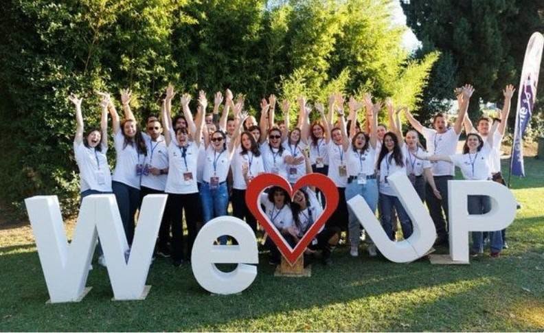 Ομάδα νέων με λευκά μπλουζάκια πανηγυρίζει μπροστά στις λέξεις "WE U…