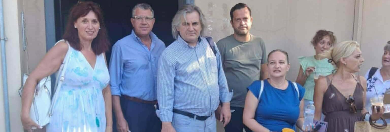 Ομάδα ανδρών και γυναικών στέκεται έξω από κτίριο με την πινακίδα I…