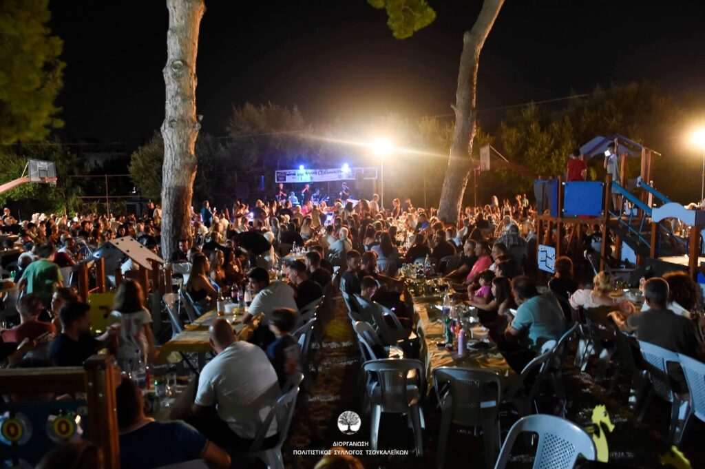 Νυχτερινό πανηγύρι με πλήθος κόσμου να διασκεδάζει σε τραπέζια, με…