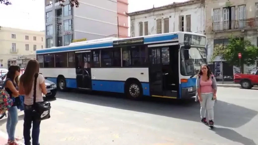 Αστικό λεωφορείο σε δρόμο της Πάτρας με πεζούς και κτίρια.