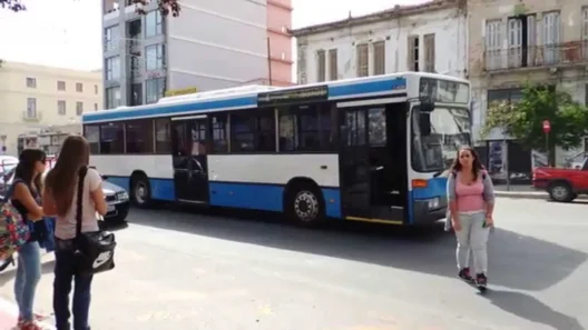 Αστικό λεωφορείο σε δρόμο της Πάτρας με πεζούς και κτίρια.