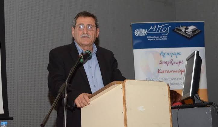 Ο Κ. Πελετίδης μιλάει από βήμα με μικρόφωνο, με πανό για τον αυτισμό…