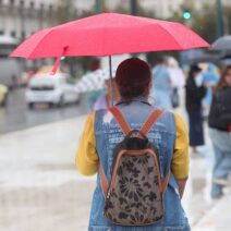 Γυναίκα με κόκκινη ομπρέλα περπατά στη βροχή σε αστικό τοπίο, με άλ…