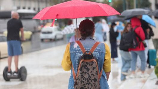 Γυναίκα με κόκκινη ομπρέλα περπατά στη βροχή σε αστικό τοπίο, με άλ…