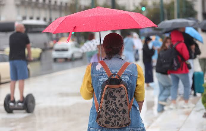 Γυναίκα με κόκκινη ομπρέλα περπατά στη βροχή σε αστικό τοπίο, με άλ…