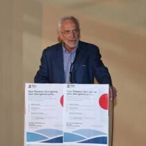 Ένας άνδρας με γκρίζα μαλλιά και γυαλιά, με μπλε σακάκι, μιλάει σε β…
