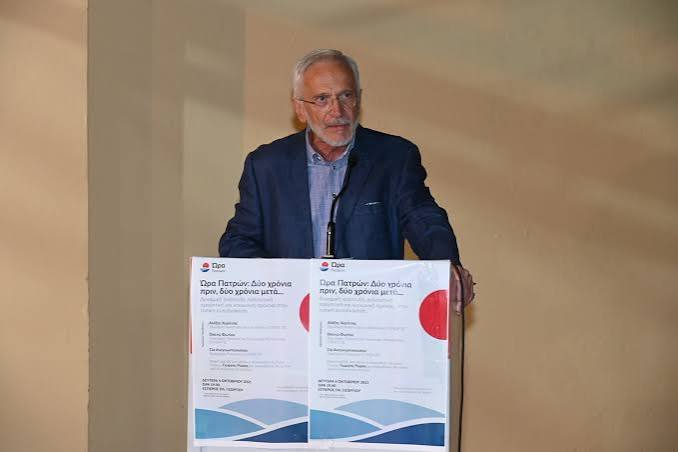 Ένας άνδρας με γκρίζα μαλλιά και γυαλιά, με μπλε σακάκι, μιλάει σε β…