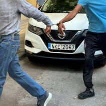 Άνδρας με χειροπέδες οδηγείται από άλλον άνδρα μπροστά σε λευκό αυ…