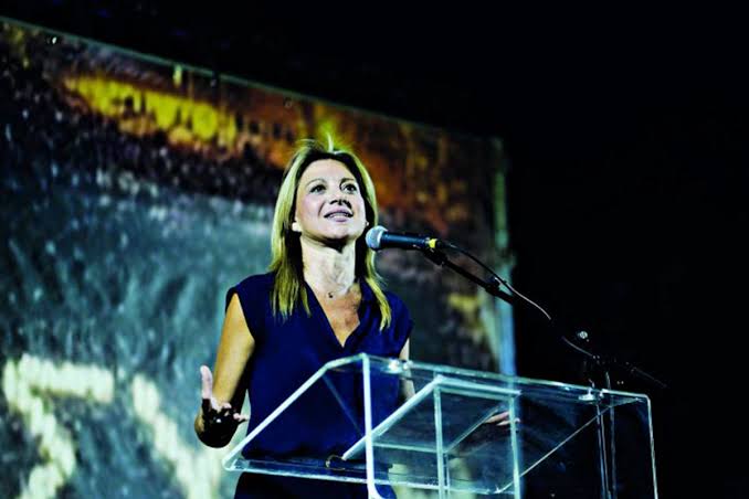 Η Μαρία Καρυστιάνου μιλάει σε εκδήλωση, πίσω της προβολή.