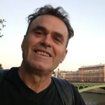 Άνδρας χαμογελά στην κάμερα, με φόντο ποτάμι, γέφυρα και κτίρια πόλ…