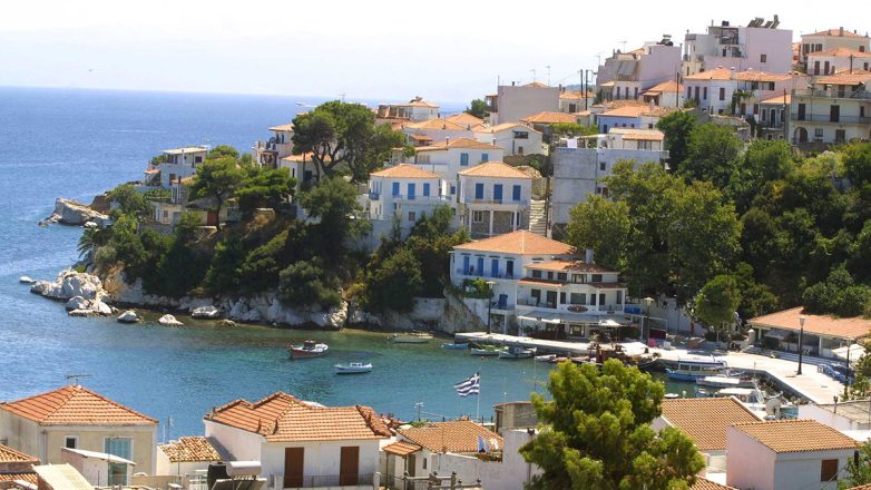 Λιμάνι Σκιάθου με λευκά σπίτια σε πλαγιά, βάρκες στη θάλασσα και ελ…