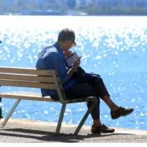 Άνδρας διαβάζει σε παγκάκι με φόντο τη λαμπερή θάλασσα, απολαμβάνο…
