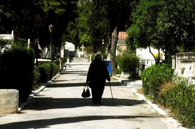 Ηλικιωμένη γυναίκα περπατά με μπαστούνι σε μονοπάτι νεκροταφείου.