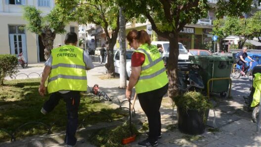 Δύο εργάτες του Δήμου Πατρέων με κίτρινα γιλέκα καθαρίζουν χόρτα σ…