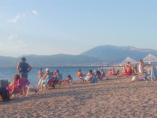 Άνθρωποι σε ηλιόλουστη παραλία με βουνά και τη Γέφυρα Ρίου-Αντιρρί…
