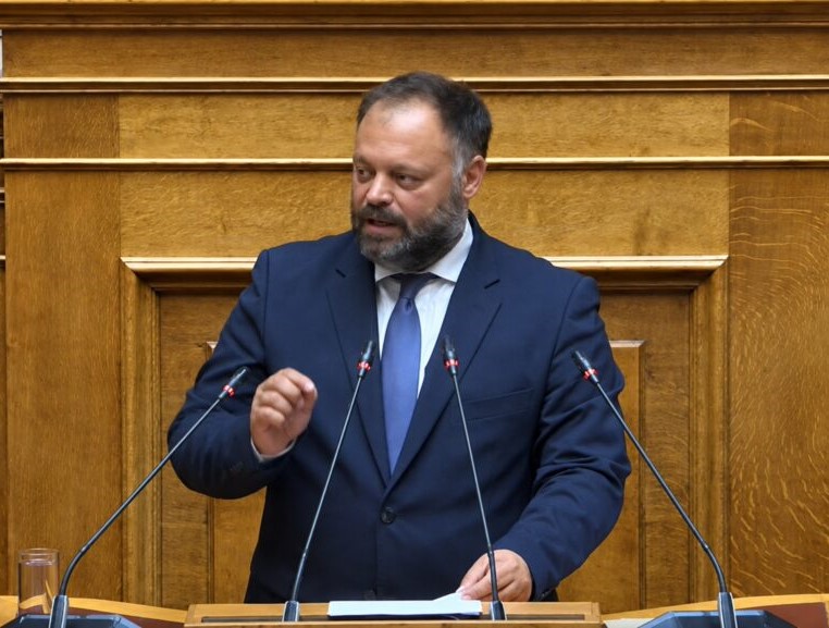 Βουλευτής μιλάει στο βήμα της Βουλής, φορώντας σκούρο κοστούμι και…